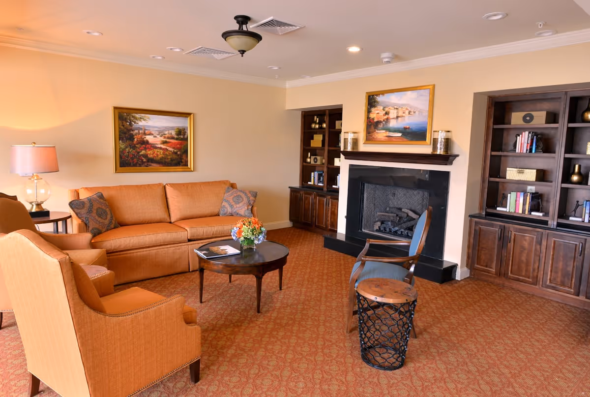 A cozy living room with an orange sofa, two matching armchairs, a wooden coffee table with a flower arrangement, a fireplace with a painting above it, and built-in wooden bookshelves filled with books and decorative items.