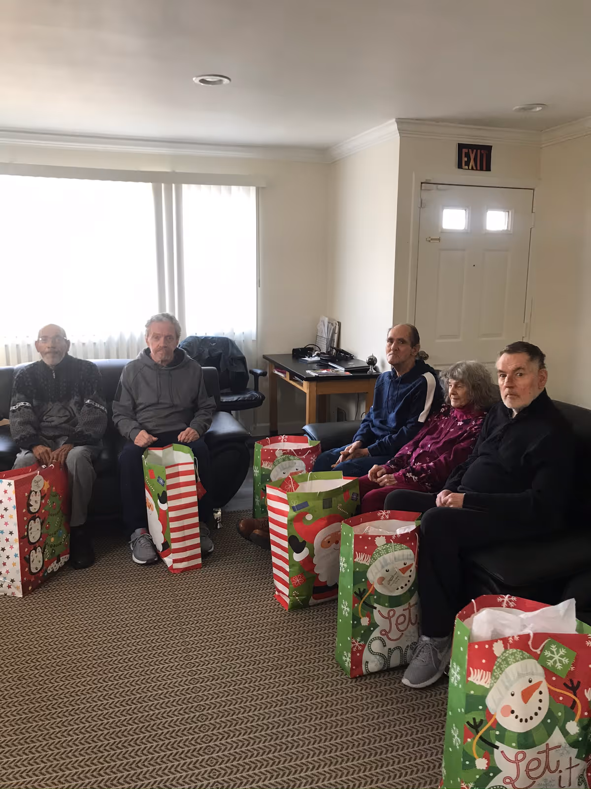 Five elderly individuals sitting on couches in a living room area, each with a large festive holiday gift bag in front of them. The room has a window with white curtains, a desk with some items on it, and an exit door with an exit sign above it.