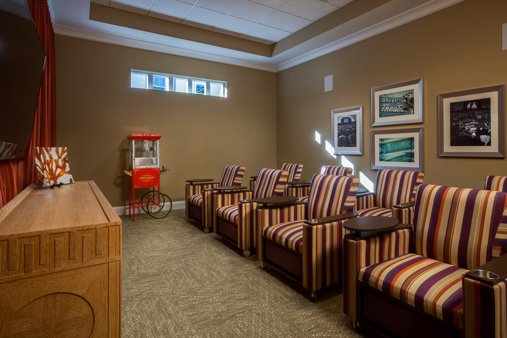 A cozy small theater room with striped armchairs arranged in rows facing a large screen. There is a vintage-style red popcorn machine against the wall, and framed pictures related to movies and theaters hang on the beige walls. A wooden cabinet is positioned near the screen.