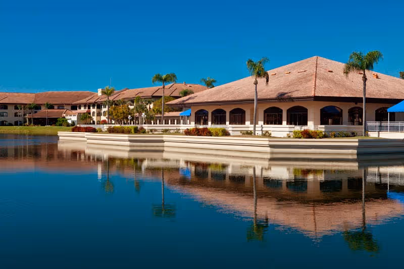 A large senior living facility building with a tiled roof and arched windows, situated beside a calm body of water reflecting the building and surrounding palm trees under a clear blue sky.