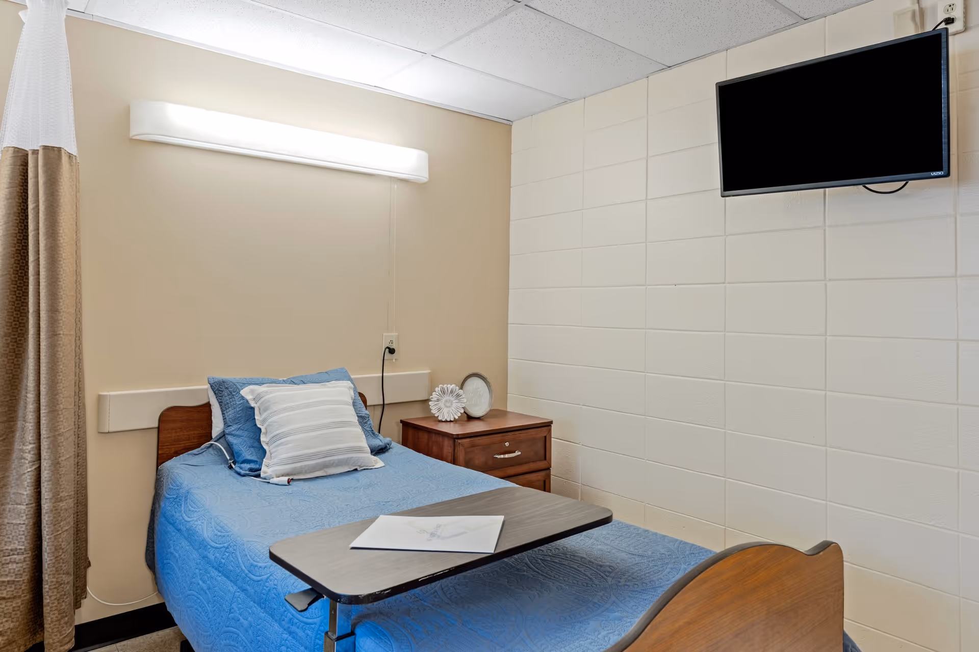 Small nursing facility patient room with a single bed covered in blue bedding, an overbed table, nightstand, wall-mounted TV and a privacy curtain.
