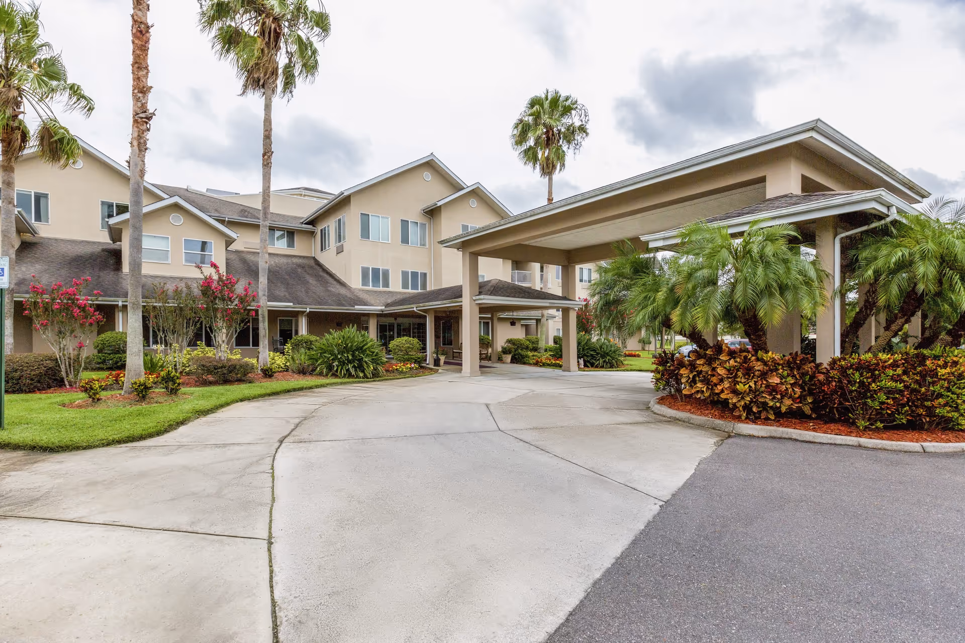 Exterior view of Azalea Park - A Provincial Senior Living Community showing a beige multi-story building with large windows, a covered driveway entrance, palm trees, and well-maintained landscaping with green bushes and flowering plants under a cloudy sky.
