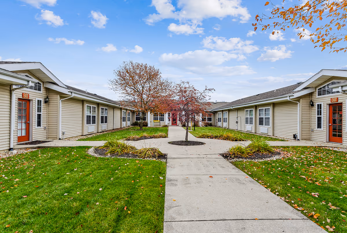 Exterior view of a single-story senior living facility with beige siding and red doors marked with exit signs. The building forms a U-shape around a landscaped courtyard with green grass, small trees, and paved walkways under a partly cloudy sky.