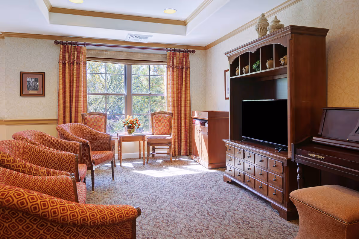 A cozy living room with patterned orange armchairs arranged around a small wooden table with a flower vase. Large windows with orange curtains let in natural light. A wooden entertainment center with a flat-screen TV and decorative items is on the right, next to a piano and a cushioned bench.
