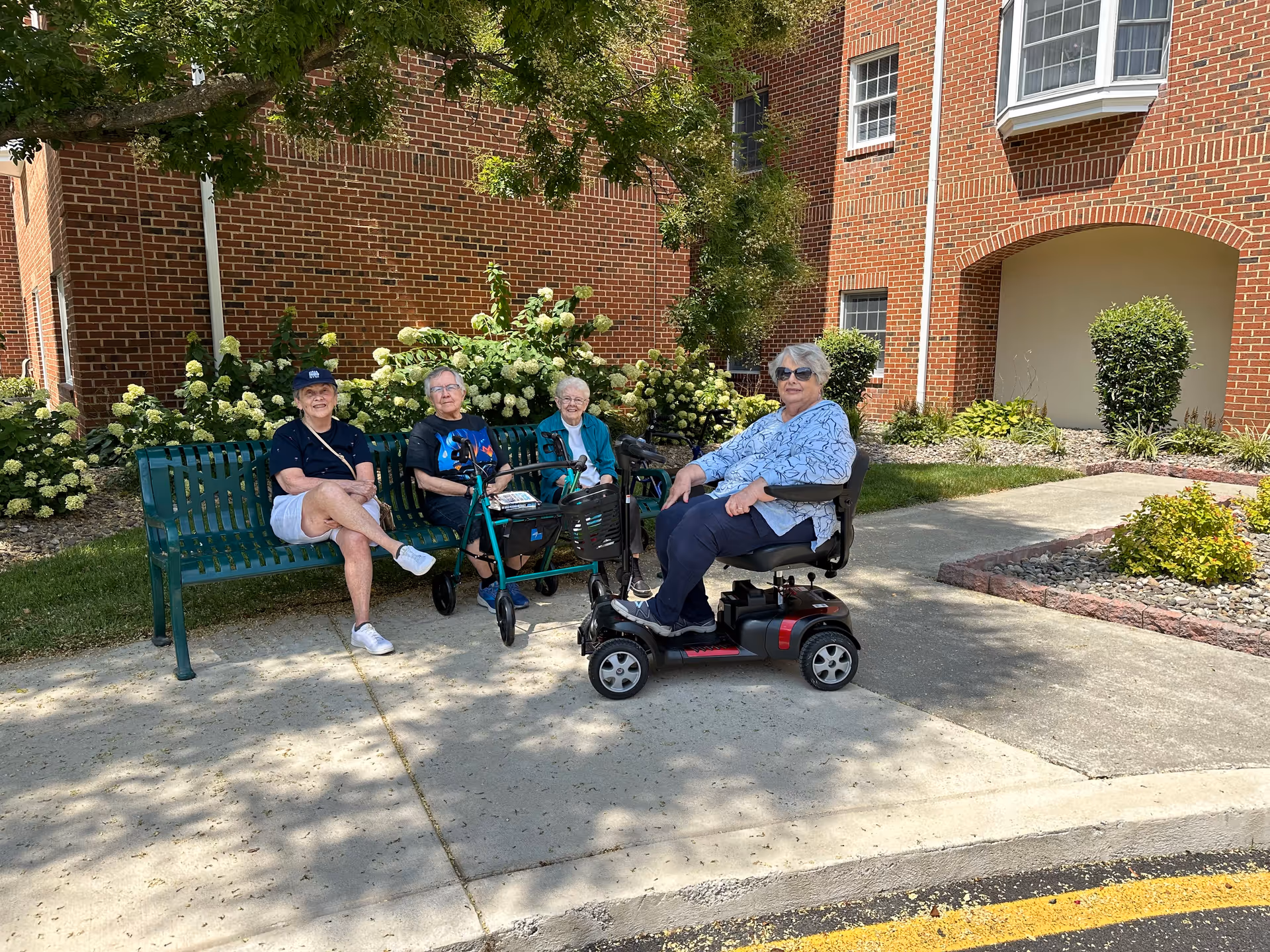 Four elderly women sitting outside near a brick building. Three women are seated on a green bench, one using a walker, and the fourth woman is sitting on a mobility scooter. There are green bushes and trees in the background, and the weather appears sunny.