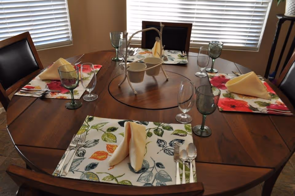 A round wooden dining table set for four with floral placemats, folded beige napkins, green and clear drinking glasses, and silverware. The table is positioned near windows with white blinds.