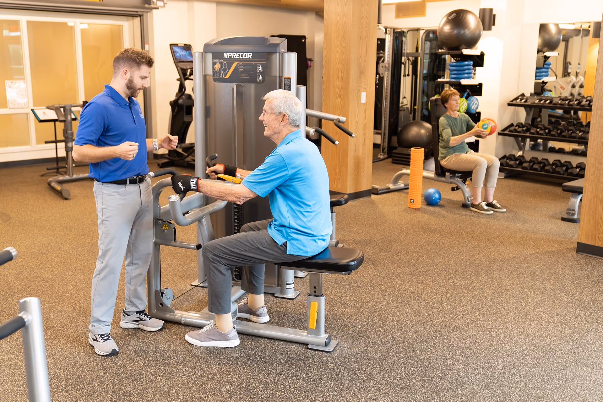 An older man uses a seated exercise machine with a trainer assisting while another older adult works with a medicine ball in the background of a fitness room.