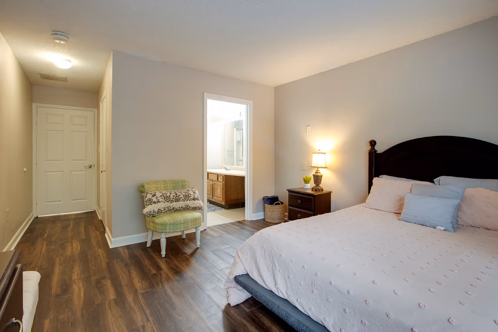 A bedroom with a large bed covered in a light pink quilt and multiple pillows. Next to the bed is a dark wooden nightstand with a lamp and a small plant. A green plaid chair with two decorative pillows is placed near the doorway leading to a bathroom. The room has wood flooring and neutral-colored walls.