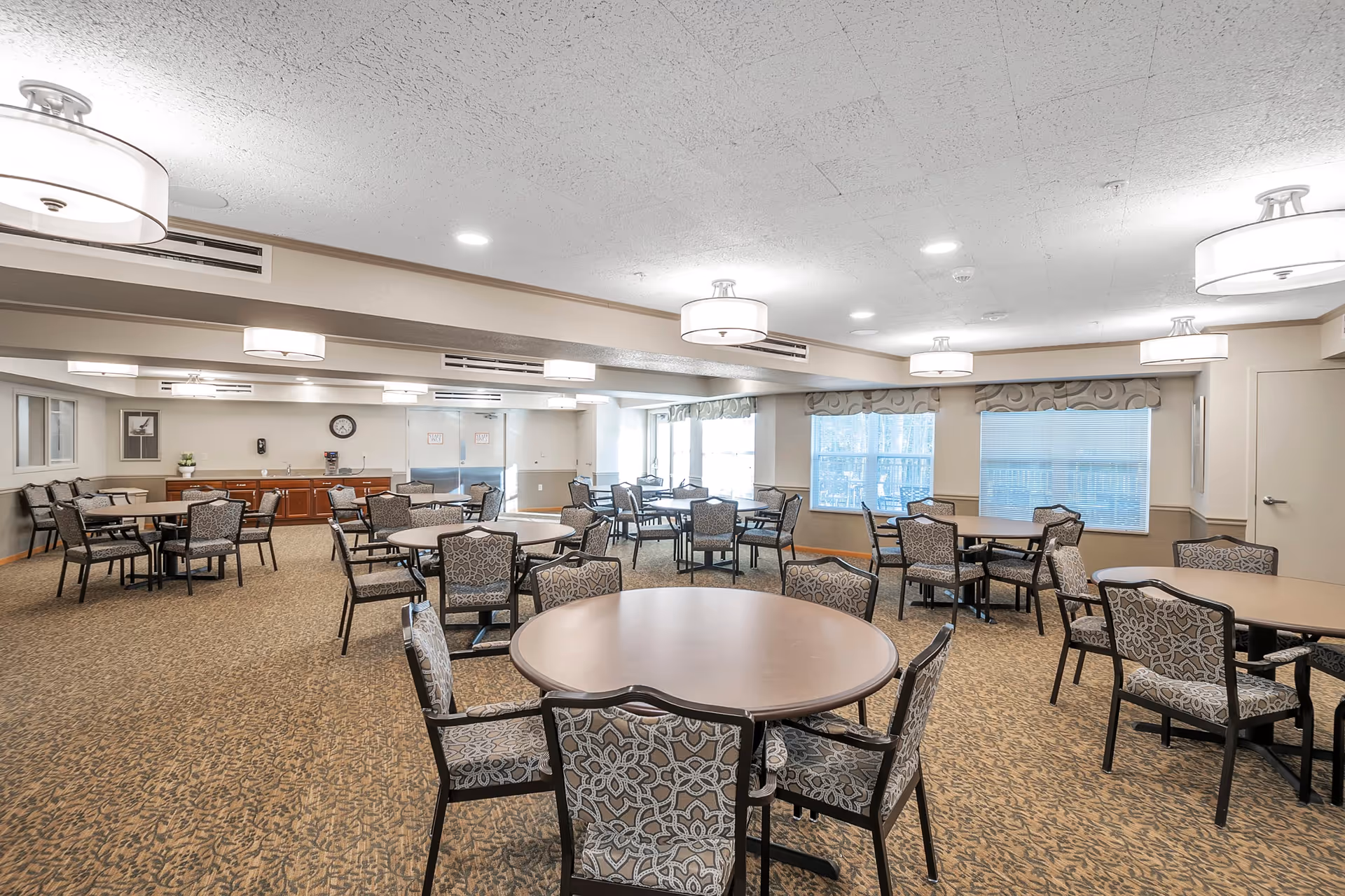 A spacious dining room in a senior living facility with multiple round tables surrounded by patterned chairs. The room is well-lit with ceiling lights and large windows with blinds, and has a carpeted floor and neutral-colored walls.