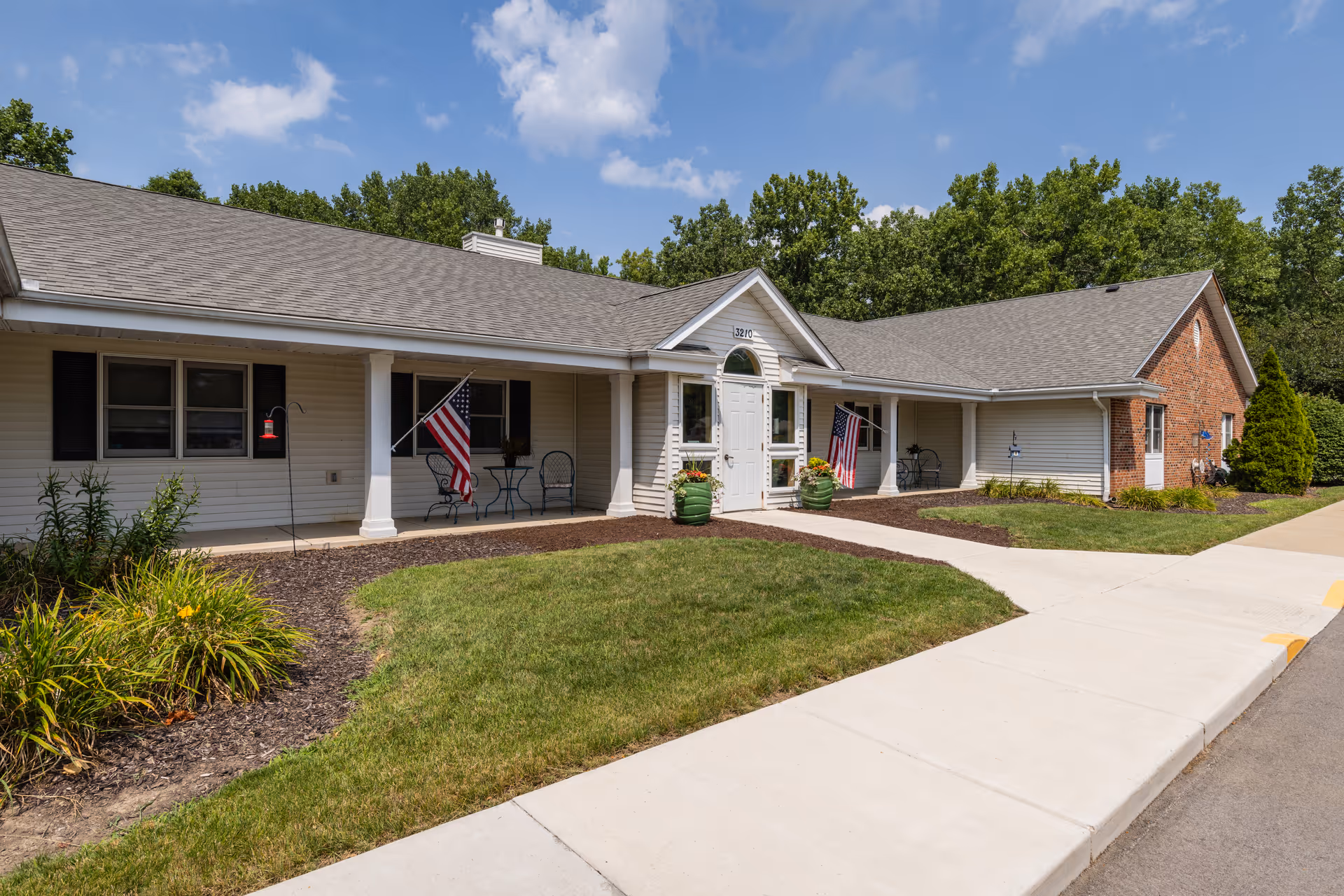 Single-story senior living building front with a covered entrance, American flags, and a paved walkway.