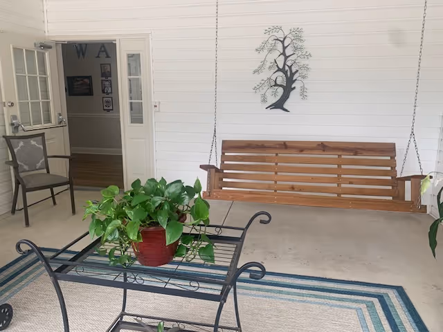A covered porch area with a wooden swing hanging from the ceiling, a metal and glass table with a potted green plant, a chair, and a decorative metal tree wall art on a white paneled wall. An open door leads to an interior hallway with framed pictures on the wall.