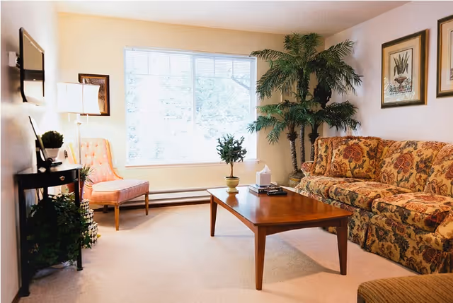 A cozy living room with a floral patterned sofa, a wooden coffee table with a small plant and tissue box on it, a pink upholstered chair near a large window with blinds, a tall green potted plant in the corner, framed botanical artwork on the wall, and a small black side table with a lamp and decorative items.