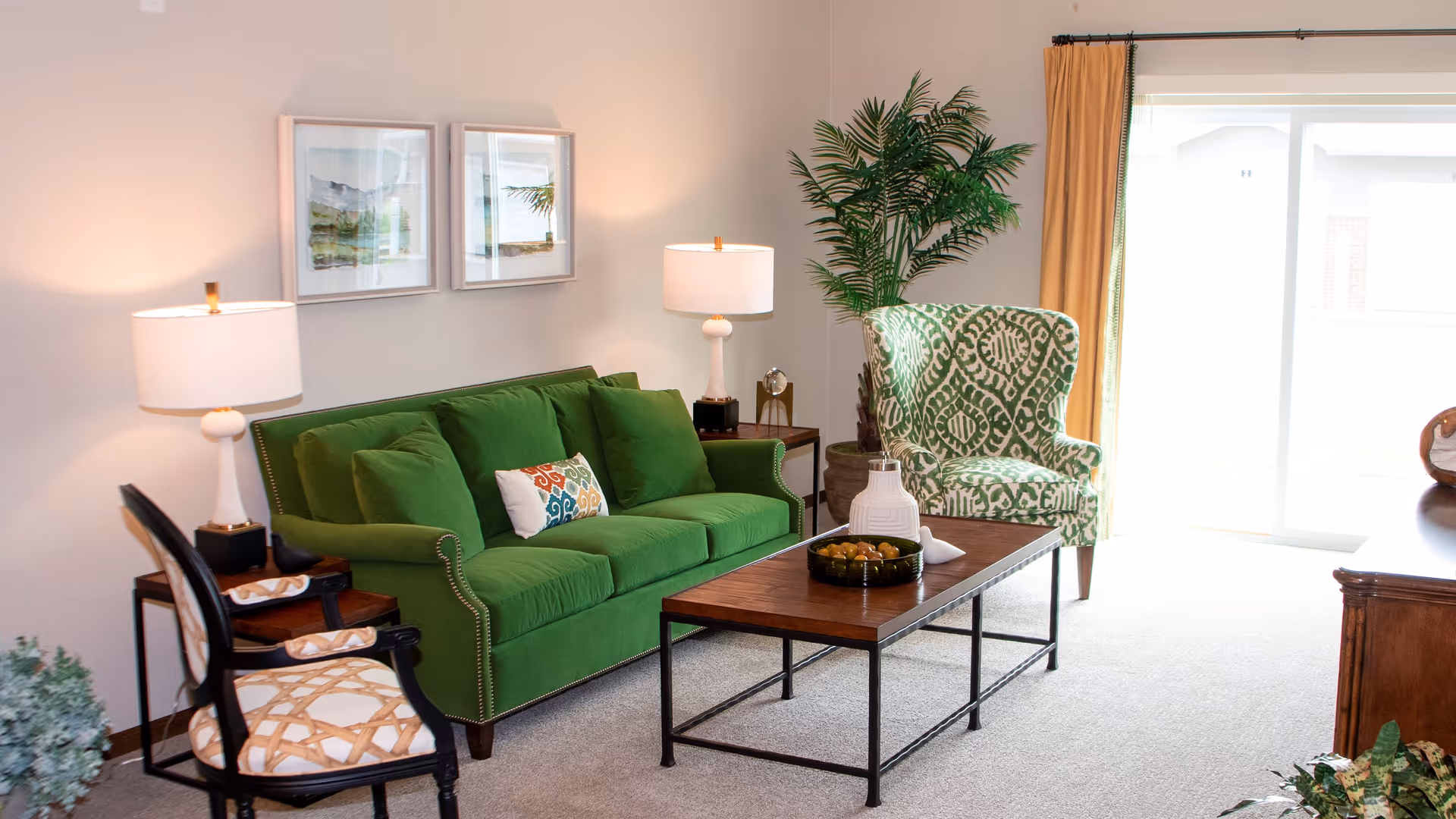 A bright living room with a green sofa adorned with multiple cushions, a patterned armchair, and a wooden coffee table with decorative items. Two table lamps with white shades sit on side tables flanking the sofa. There are two framed pictures on the wall above the sofa and a large potted plant near a sliding glass door with beige curtains, allowing natural light to fill the room.