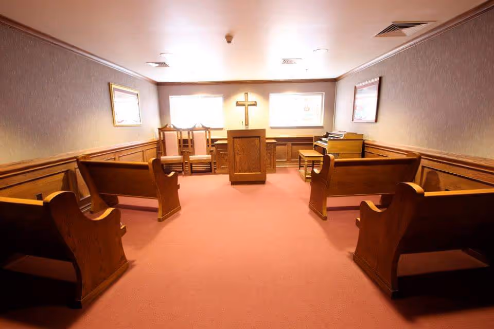 Interior view of a small chapel or prayer room with wooden pews arranged on both sides, a wooden podium with a cross on the wall behind it, two windows letting in natural light, framed pictures on the walls, and a piano on the right side.