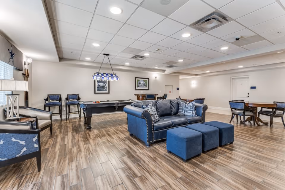 A spacious senior living common area with a blue leather couch, three blue ottomans, several chairs, a pool table with a hanging light fixture above it, and multiple tables with chairs. The room has wood-look flooring, white walls, and a drop ceiling with recessed lighting.