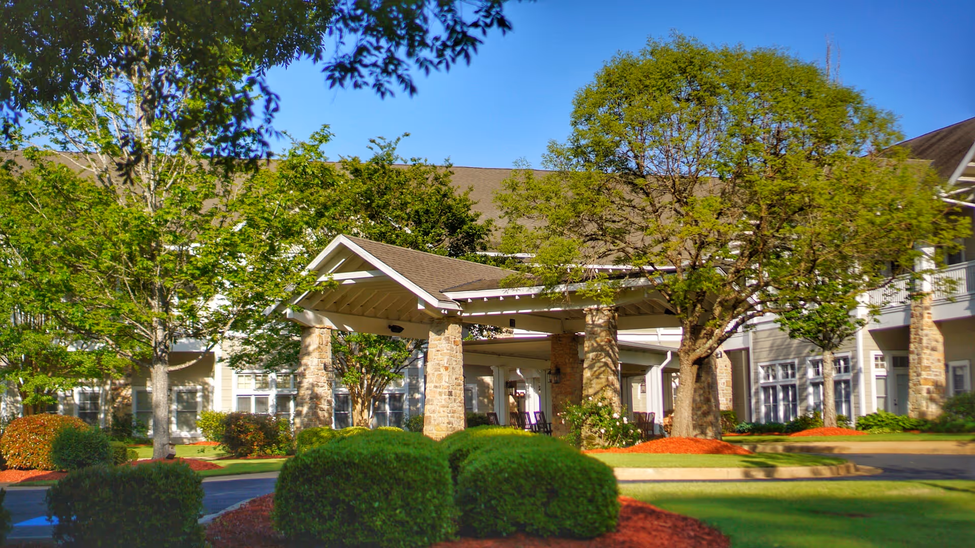 Exterior view of Vernon Woods facility showing a covered entrance supported by stone pillars, surrounded by green trees, bushes, and well-maintained landscaping under a clear blue sky.
