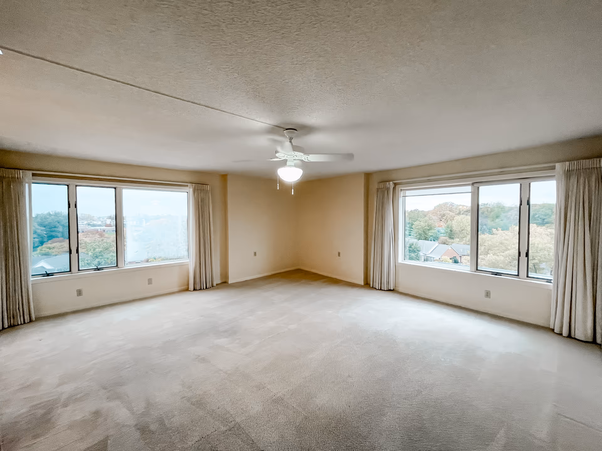 Spacious empty carpeted living room with two large windows, cream-colored walls, drapes, and a ceiling fan.