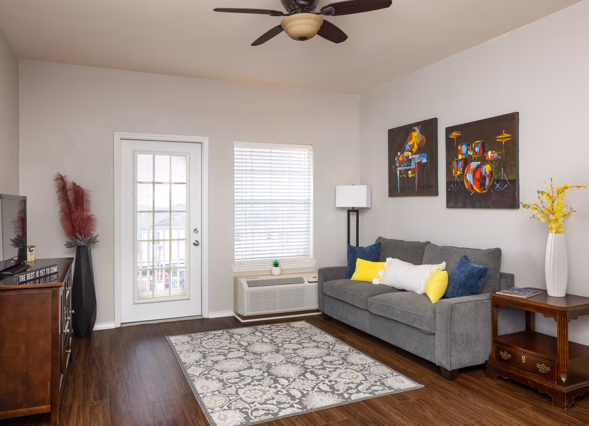 A cozy living room with a gray sofa adorned with yellow and blue pillows, a patterned area rug on wooden flooring, a floor lamp, and two colorful paintings of musical instruments on the wall. There is a window with blinds and a glass door leading outside. A wooden side table with a white vase holding yellow flowers and a wooden cabinet with decorative items are also visible.