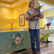 A man wearing a cap, glasses, and a beige shirt stands indoors holding a small kangaroo. Behind him is a table covered with a green cloth featuring a logo with the text 'LOWMAN WILDLIFE SANCTUARY'. The room has yellow walls, a lamp, framed artwork, and a window with a small table and decorations.
