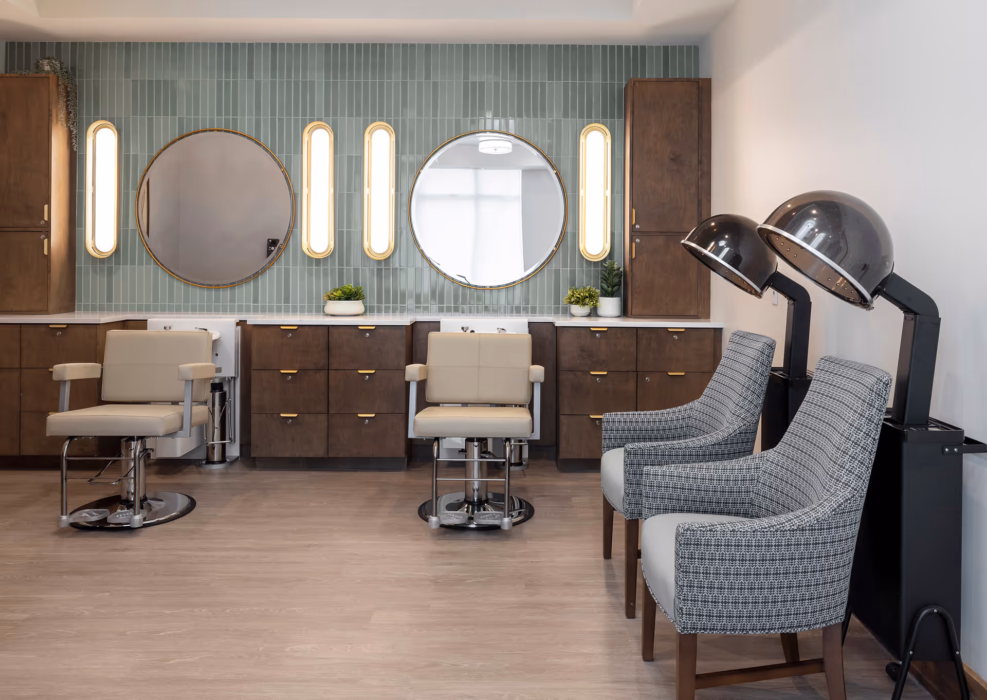 Interior view of a salon area with two beige salon chairs in front of a wall with green vertical tiles, two round mirrors, and vertical light fixtures. There are wooden cabinets below the mirrors and two gray patterned chairs with hair dryers on the right side.