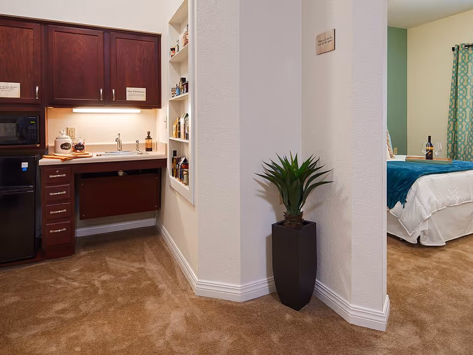 Interior view of a senior living facility showing a small kitchenette with dark wood cabinets, a microwave, mini fridge, and a sink. Adjacent to the kitchenette is a corner with a tall black planter holding a green plant. In the background, a bedroom area is partially visible with a bed covered in white bedding and a teal throw, a window with teal patterned curtains, and a tray with a wine bottle and glass on the bed.