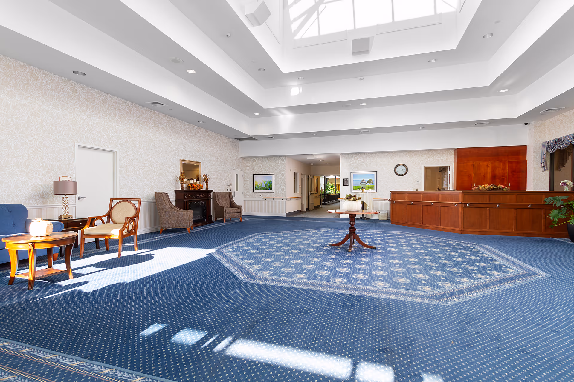 A spacious, well-lit lobby area with a large blue patterned carpet, a round wooden table with a floral centerpiece in the center, several chairs and small tables along the left wall, a wooden reception desk on the right, and framed artwork and a clock on the far wall. The ceiling features a large skylight allowing natural light to fill the room.