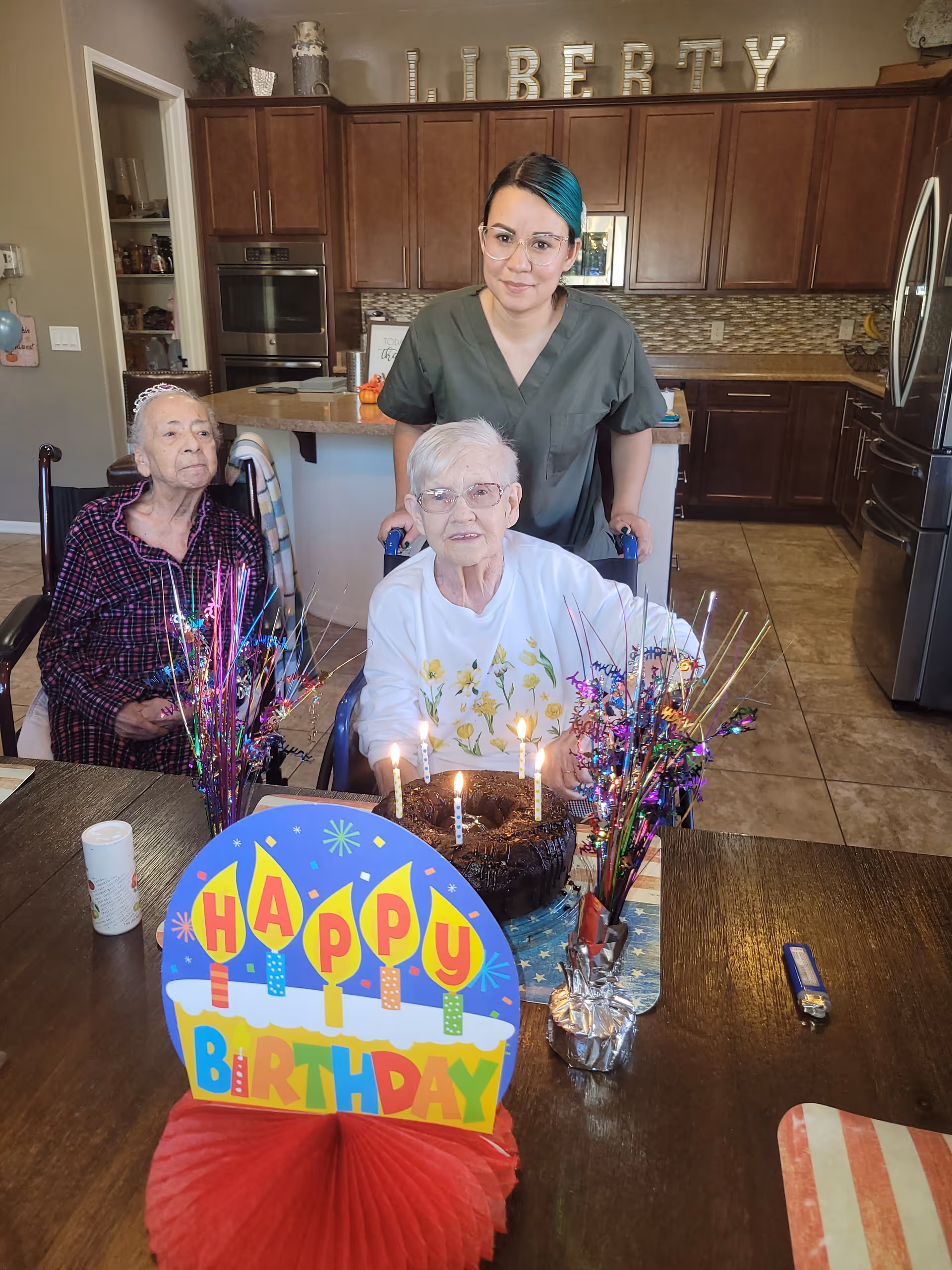 A caregiver stands behind two elderly women seated at a table with a lit birthday cake and colorful decorations in a kitchen with 'LIBERTY' letters on the cabinets.