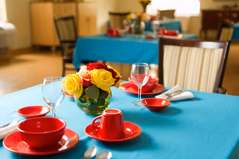 A dining table set with a turquoise tablecloth, red dishes including bowls, plates, and cups, silverware, and wine glasses. A small vase with yellow and red roses is placed in the center of the table. In the background, other similarly set tables and chairs are visible in a well-lit dining room.