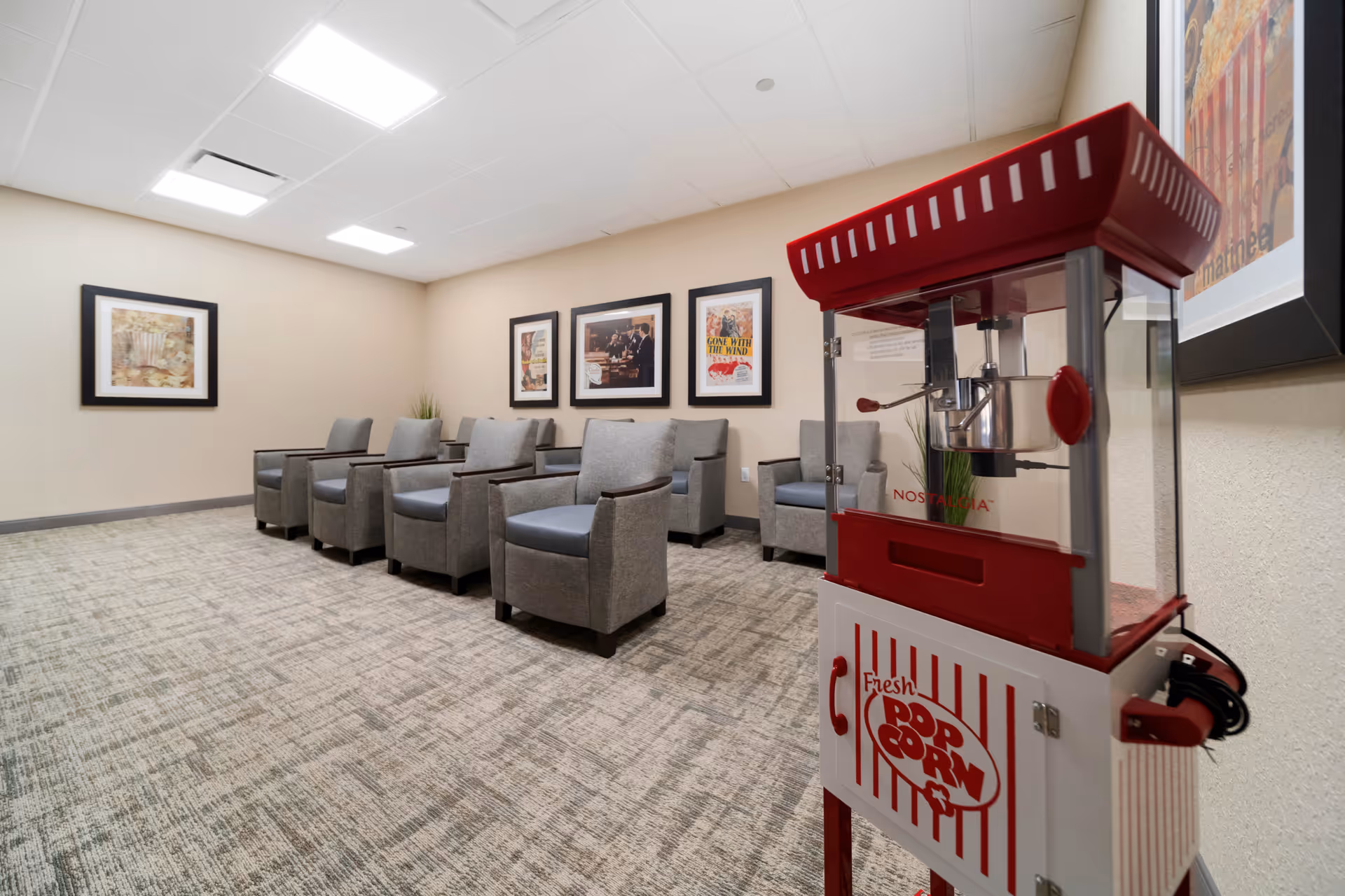 A cozy room with six gray armchairs arranged in two rows facing the same direction. The walls are decorated with framed pictures, and there is a red and white popcorn machine in the foreground on the right side. The room has a carpeted floor and ceiling lights.