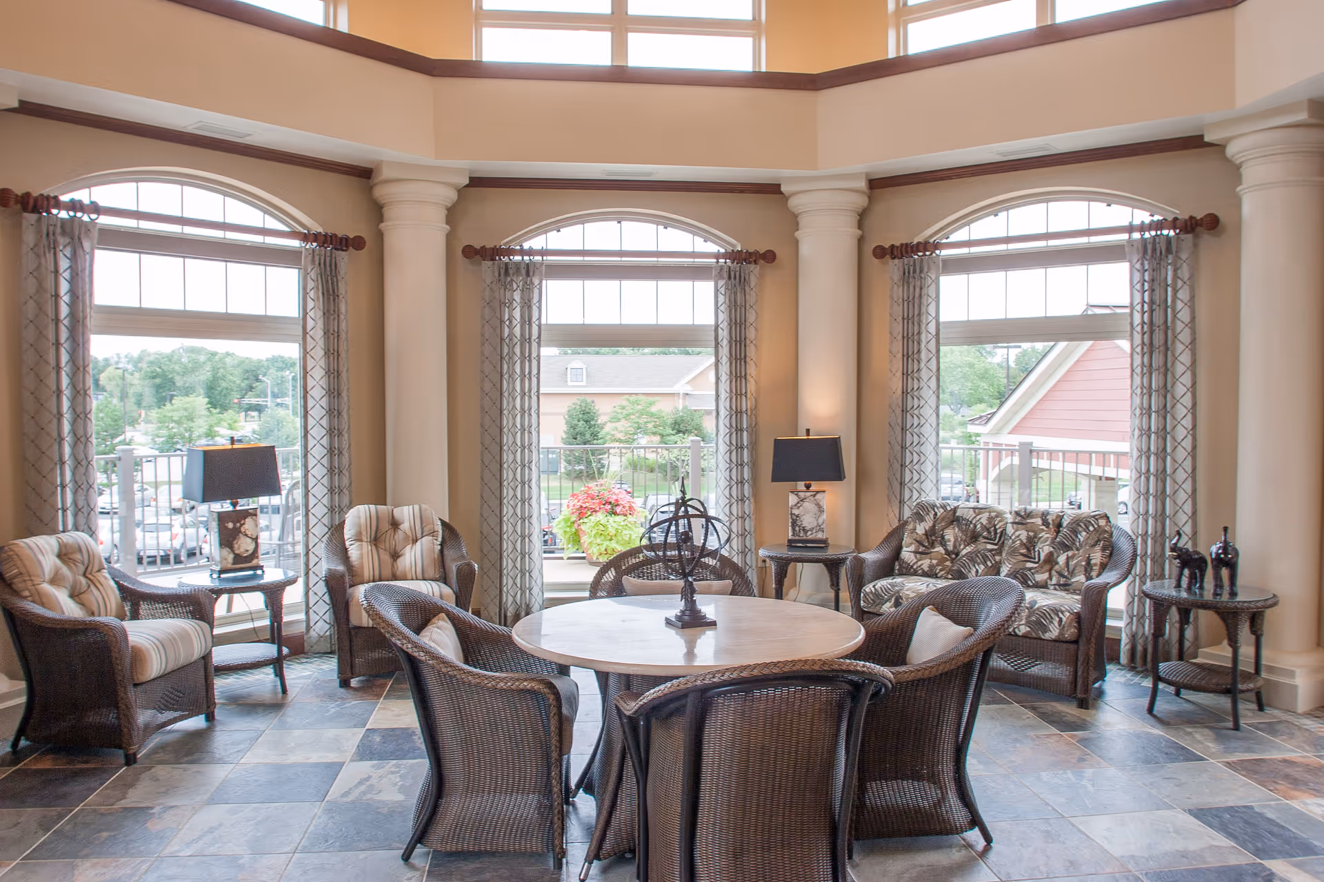 A bright and spacious sitting area with large arched windows letting in natural light. The room features a round wooden table surrounded by six wicker chairs with cushions. There are additional wicker armchairs and a loveseat with patterned cushions, side tables with lamps, and decorative items. The floor is tiled with a checkered pattern, and the walls have large columns and a high ceiling with upper windows.