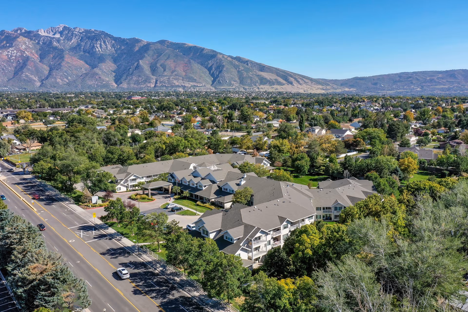 Aerial view of Solstice Senior Living at Sandy facility surrounded by trees and residential neighborhoods, with mountains in the background under a clear blue sky.