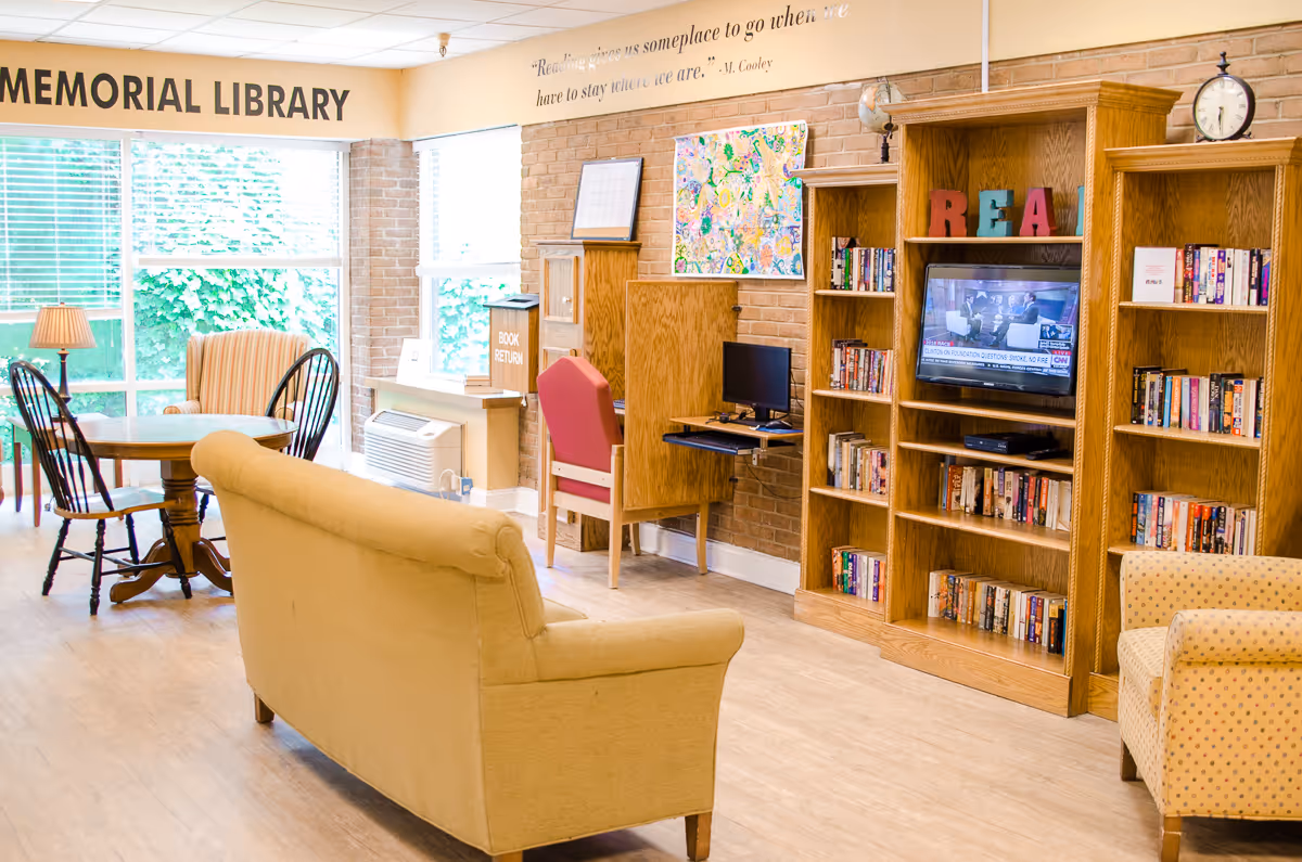 A bright and cozy library room with large windows letting in natural light. The room features a beige couch, a round wooden table with four chairs, a computer desk with a red chair, and wooden bookshelves filled with books. A TV is mounted in one of the bookshelves, and colorful letters spelling 'READ' are displayed on top. The wall has a quote and the words 'MEMORIAL LIBRARY' are prominently displayed near the ceiling.
