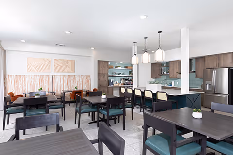 A modern dining room with multiple dark wood tables and chairs with green cushions. There is a kitchen area in the background with a large island, bar stools, pendant lights, and stainless steel appliances. The walls are white with decorative artwork and a patterned accent wall behind a bench seating area.