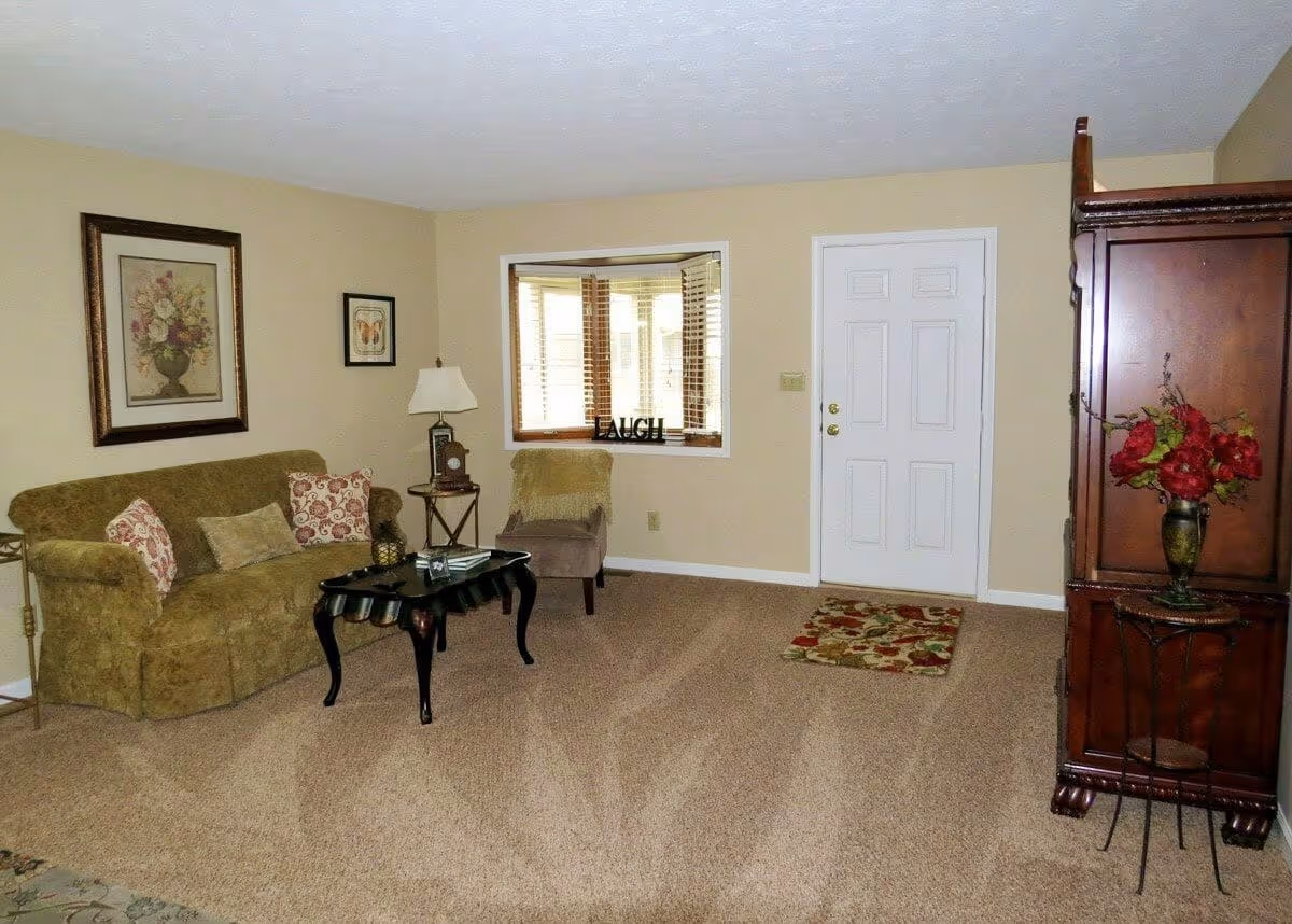 A cozy living room with beige walls and carpet. There is a green sofa with patterned cushions on the left, a small black coffee table in front of it, and a beige armchair near a bay window with wooden blinds. The word 'LAUGH' is displayed on the window sill. A white door is on the right side of the room, next to a wooden cabinet with a vase of red flowers on a small round table.