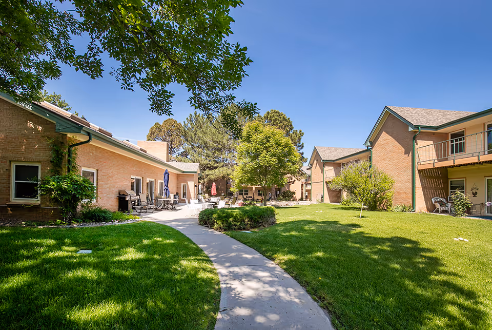 Outdoor view of a senior living facility courtyard with a curved concrete pathway, green grass, trees, and shrubs. The buildings are made of light brown brick with green trim and have patios and balconies with outdoor furniture. The sky is clear and blue.