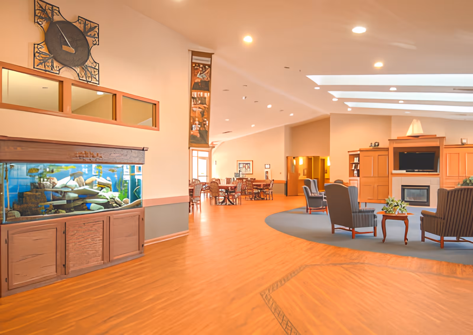 A spacious senior living facility common area with wooden flooring, a large fish tank built into a wooden cabinet, a decorative wall clock above the tank, and a seating area with armchairs and a small table near a fireplace and TV. In the background, there are additional tables and chairs for dining or socializing, with soft lighting and a high ceiling with skylights.