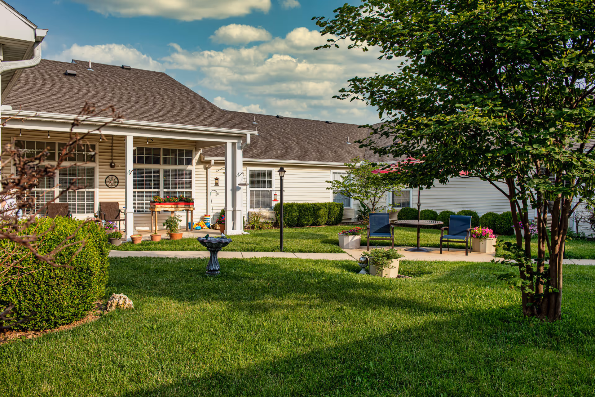 A well-maintained outdoor garden area at Alderbrook Village featuring green grass, trimmed bushes, a birdbath, and a patio with two blue chairs and a round table under a red umbrella. The background shows beige residential buildings with large windows and a covered porch with potted plants and chairs. The sky is partly cloudy with blue patches.