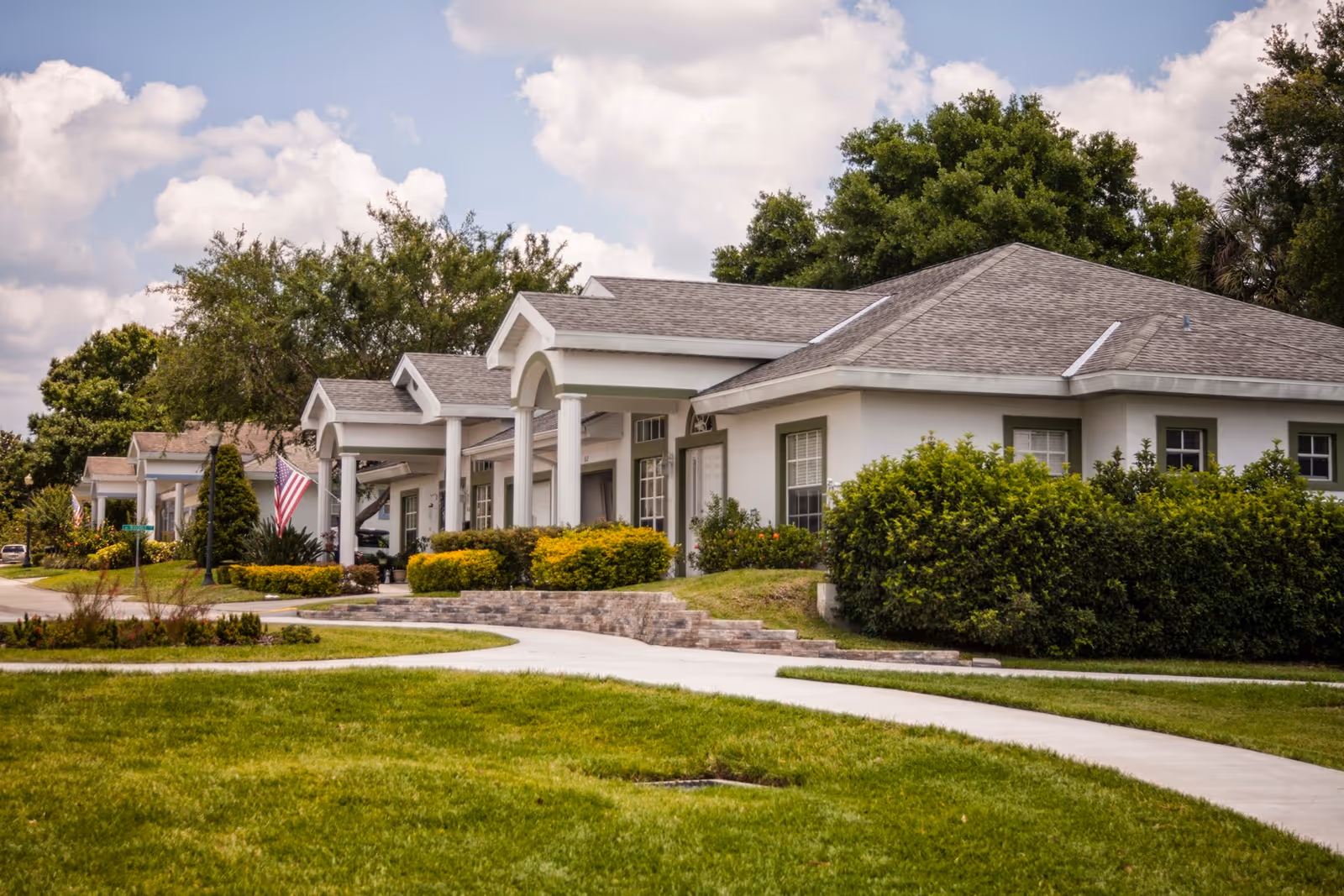 Single-story senior living buildings with columned porches, manicured landscaping, and American flags along a curved sidewalk.