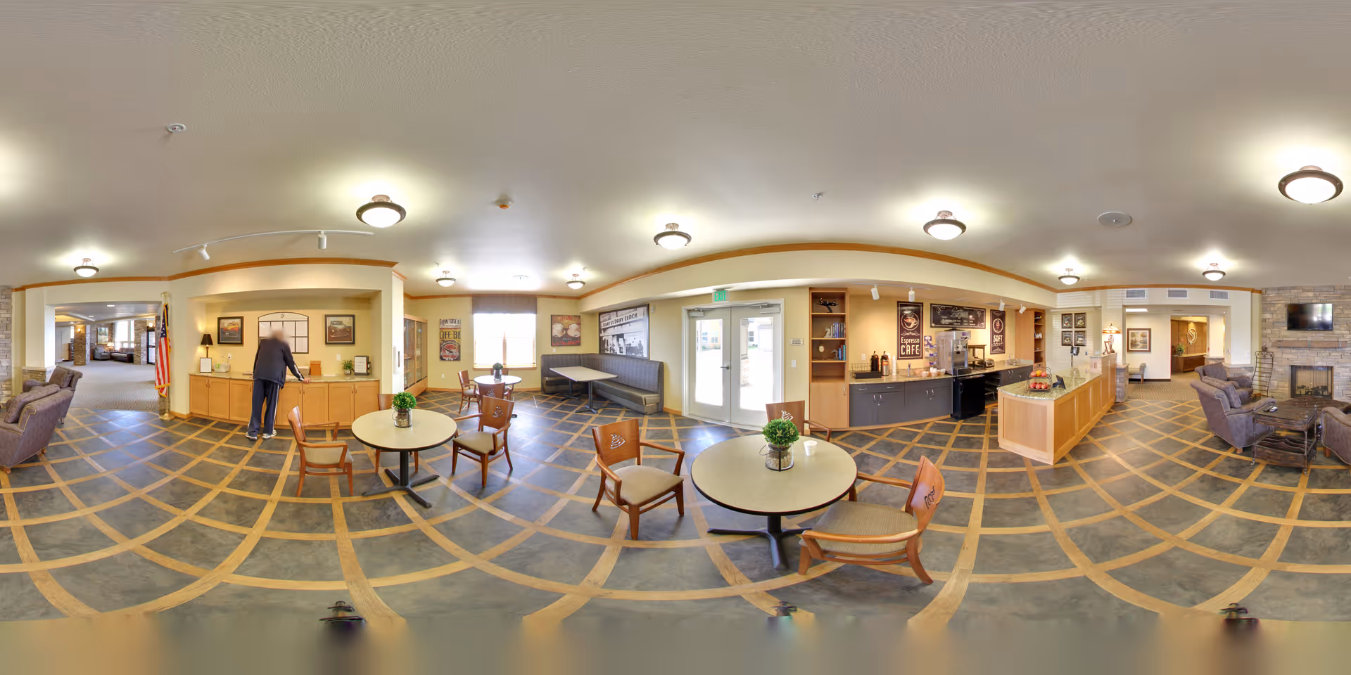 A spacious senior living facility common area with multiple round tables and chairs, a small café counter with coffee machines and signage, a seating area with armchairs near a stone fireplace, and a person standing at a counter on the left side. The room has a patterned floor, warm lighting, and large windows allowing natural light.