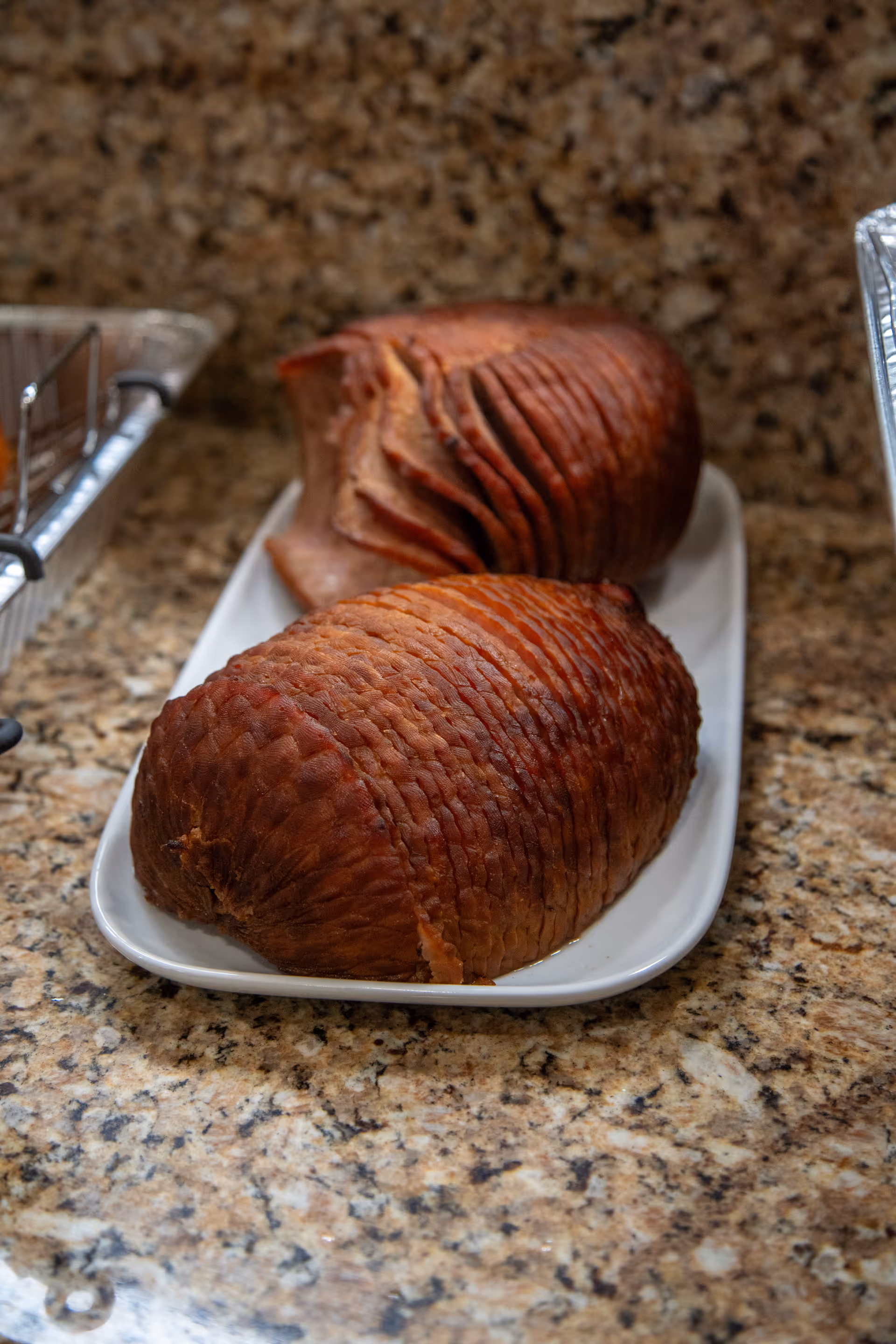 Two sliced glazed hams on a white platter set on a speckled granite countertop.