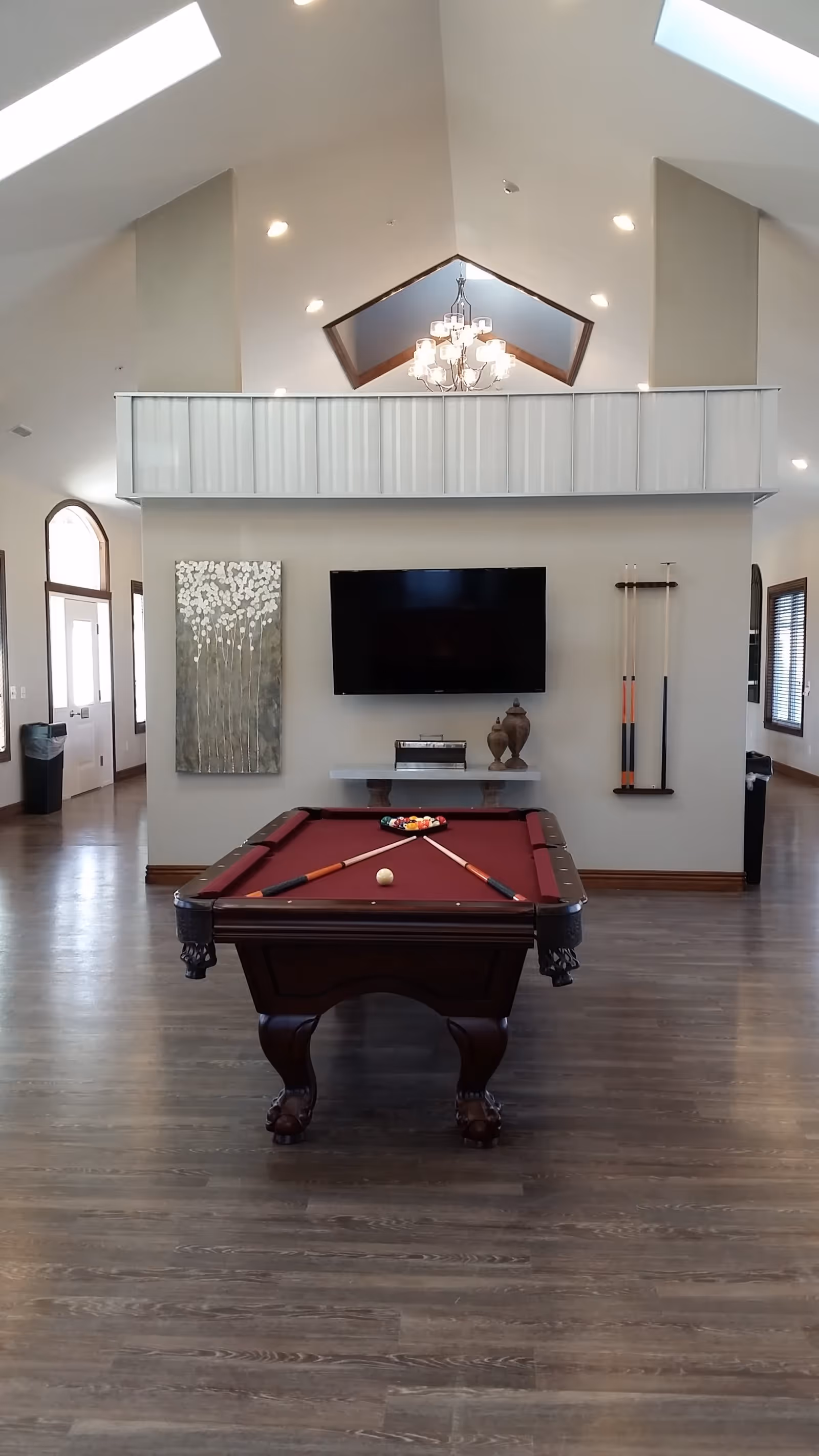Spacious common room with a burgundy pool table centered beneath a vaulted ceiling, wall-mounted TV, cue rack and chandelier.