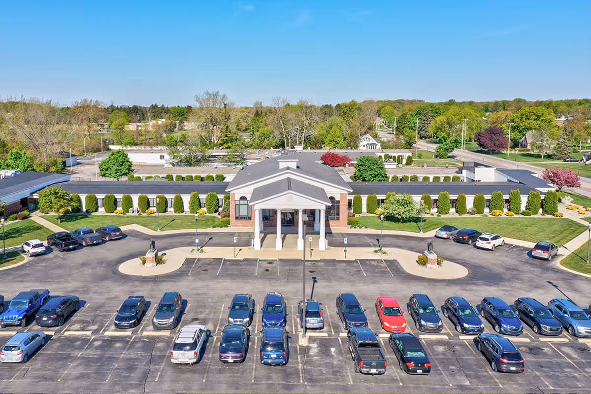 Aerial view of Carriage House Nursing and Rehab building with a large parking lot filled with cars in front. The building has a central entrance with columns and is surrounded by well-maintained landscaping including bushes and trees. The sky is clear and blue.