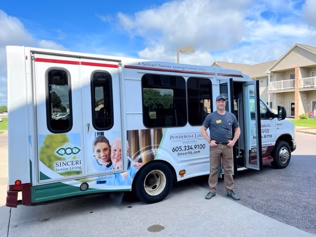 A Ponderosa Lodge Senior Living shuttle bus parked outside with a smiling staff member standing by the open door.