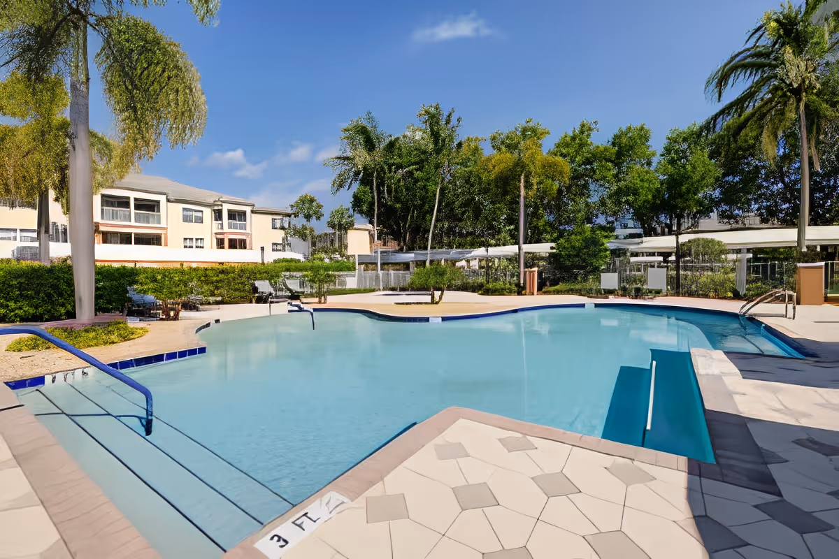 Outdoor swimming pool with steps and handrails, surrounded by a tiled deck, palm trees, and greenery. In the background, there are multi-story residential buildings under a clear blue sky.
