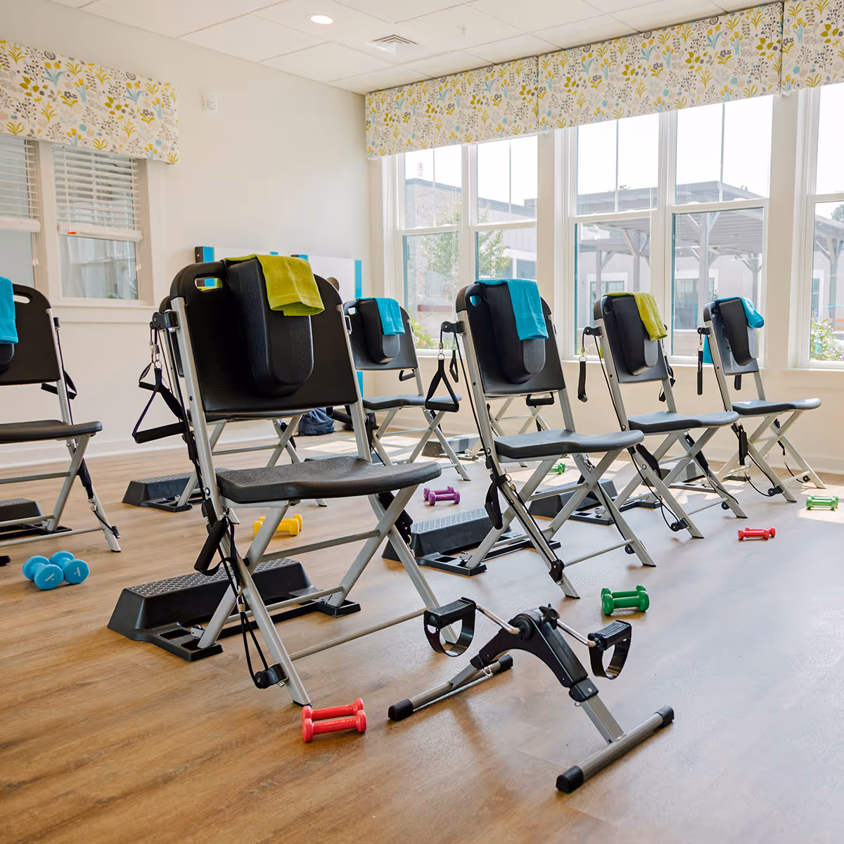 A bright exercise room with several black folding exercise chairs equipped with resistance bands and small towels draped over the backrests. Colorful small dumbbells and step platforms are scattered on the wooden floor. Large windows with floral valances allow natural light to fill the room.