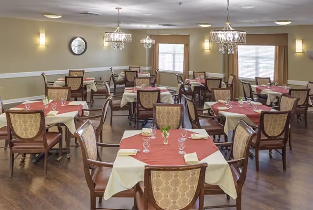 Dining room with multiple tables covered with red and white tablecloths, each set with glassware, napkins, and small flower arrangements. The room has wooden chairs with patterned upholstery, hardwood floors, wall-mounted lights, and windows with beige curtains allowing natural light.