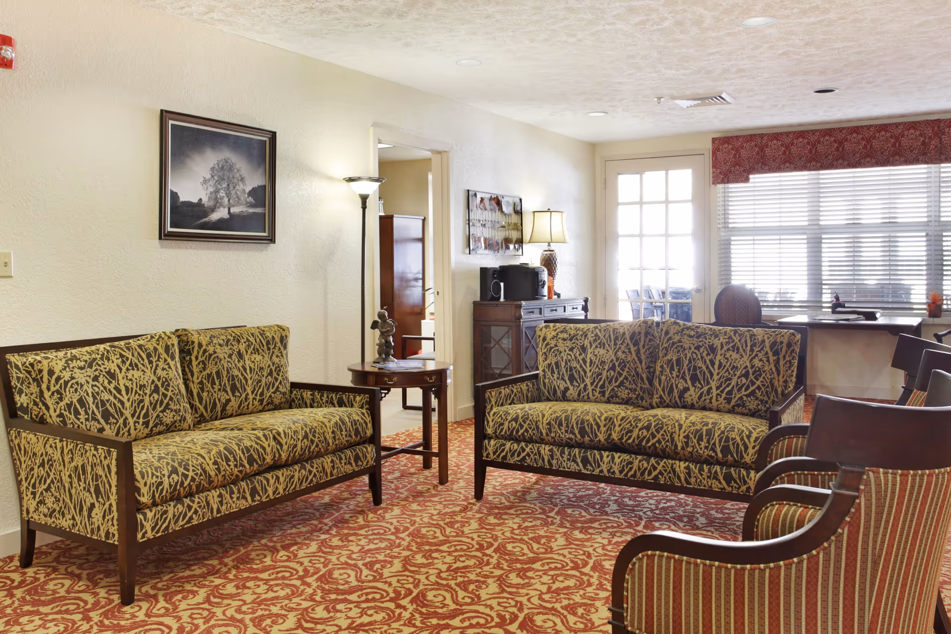 A cozy living room area with two patterned sofas facing each other, a round wooden side table with a small sculpture, a floor lamp, and a framed picture of a tree on the wall. The room has a patterned red carpet, a window with blinds and a red valance, and a door leading to another room with additional furniture visible.