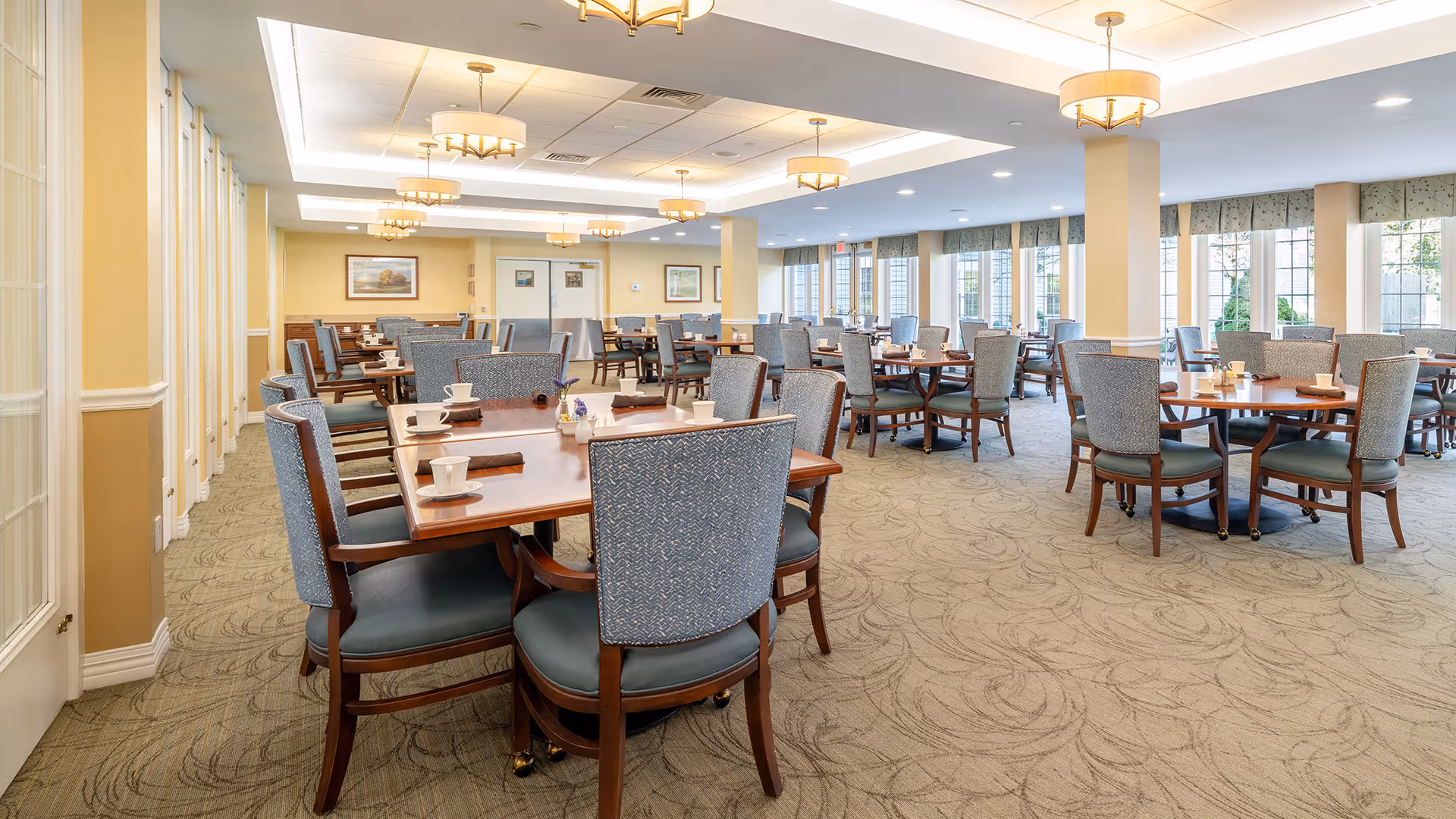 A spacious dining room with multiple wooden tables and cushioned chairs arranged neatly. Each table is set with cups, saucers, and napkins. The room has large windows allowing natural light to brighten the space, and ceiling lights with round lampshades. The walls are painted in light colors with framed artwork hanging.