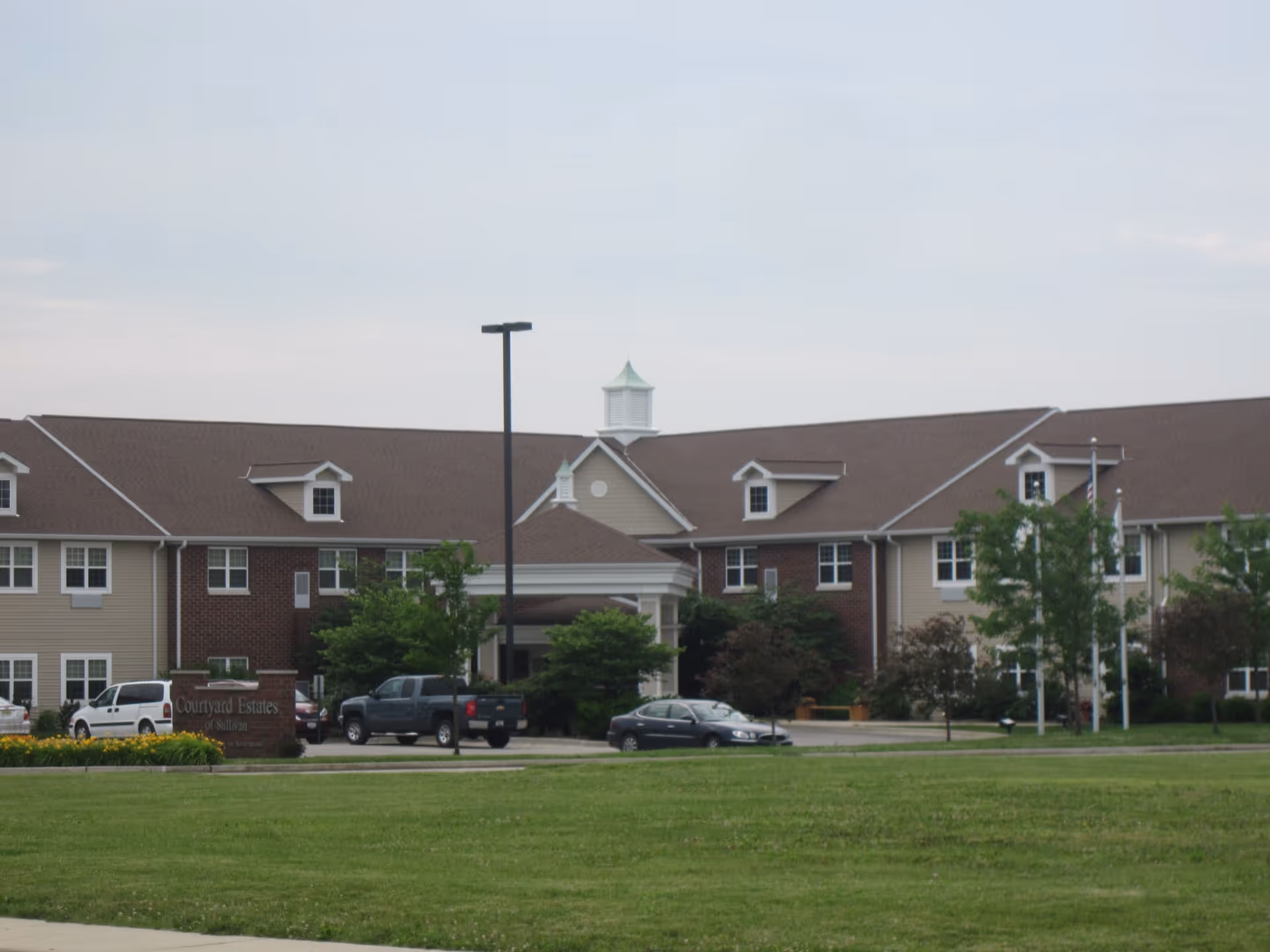 Exterior view of a two-story senior living facility building with a brown roof, beige and brick walls, several windows, and a small cupola on top. There are a few vehicles parked in front and a green lawn in the foreground under a cloudy sky.