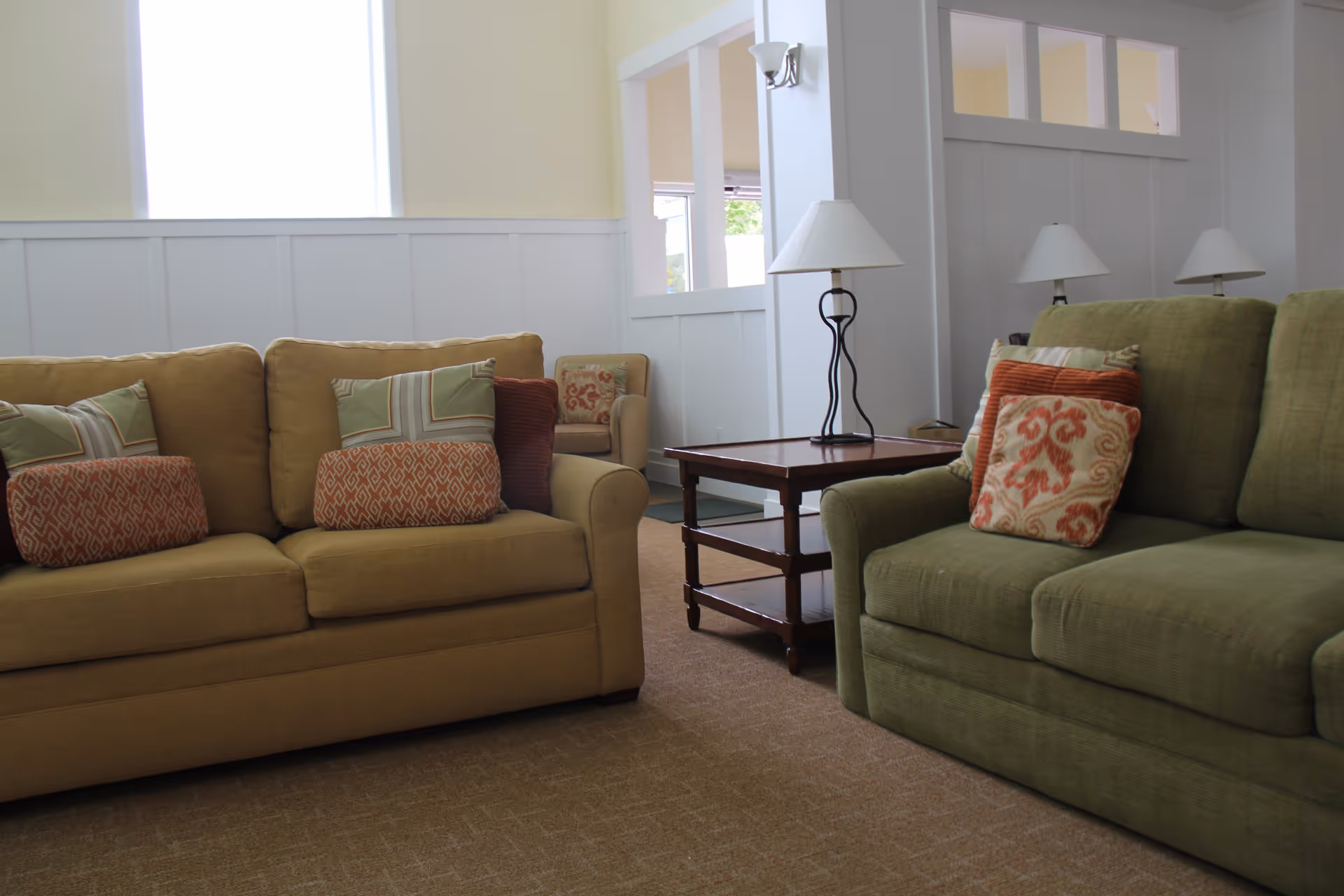 A cozy living room area with two sofas, one beige and one green, each adorned with patterned throw pillows. Between the sofas is a wooden side table with a lamp on it. The room has light-colored walls and carpeted flooring, with a window letting in natural light.